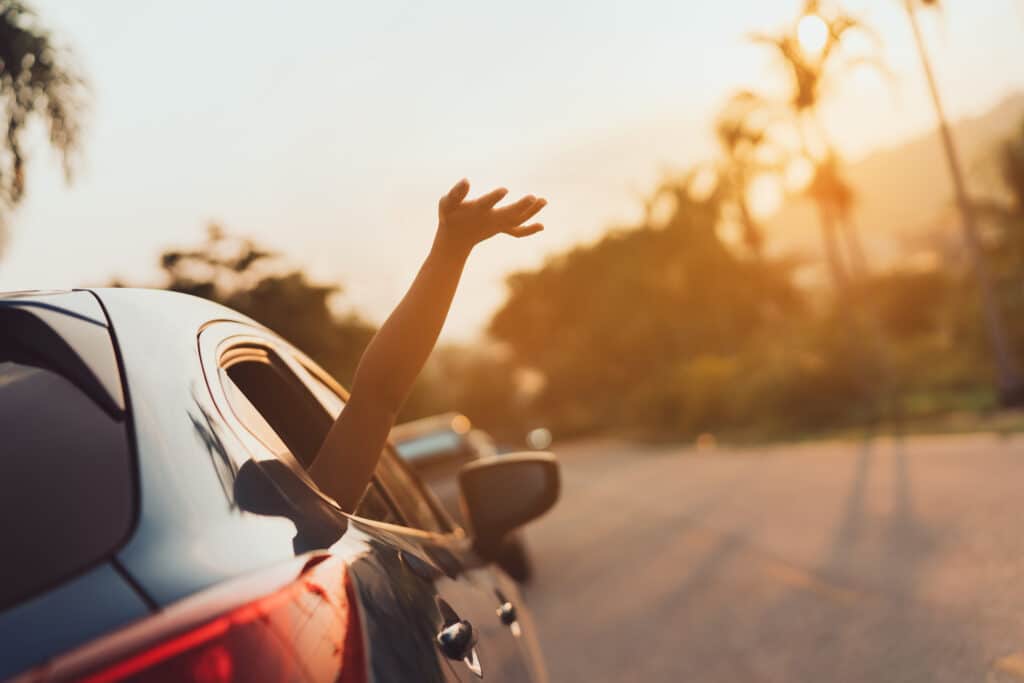Main tendue hors d'une fenêtre de voiture au coucher du soleil, avec palmiers flous. Évoque le voyage, la liberté et l'été.
