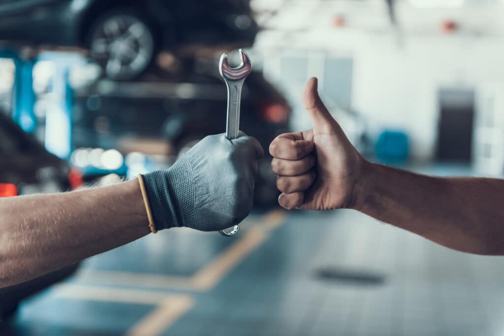 Main gantée tenant une clé de 18 et main levant le pouce dans un garage auto, signifiant satisfaction après réparation.
