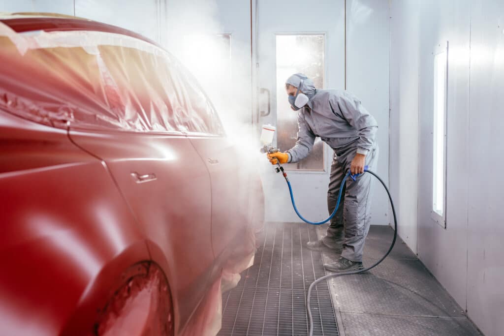 Carrossier en combinaison intégrale et masque, peignant une voiture rouge avec un pistolet dans une cabine de peinture. Brouillard visible.