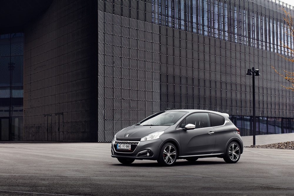 Une Peugeot 208 grise, 3 portes, avec jantes sportives, sur un parking moderne. Fond: bâtiment sombre texturé. Plaque WE 974ME.