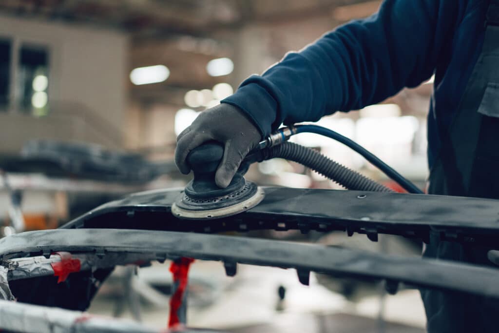 Préparation carrosserie : ponçage de pare-chocs professionnel Main gantée ponçant un pare-chocs de voiture gris avec une ponceuse orbitale pneumatique dans un atelier de carrosserie.