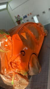 Carrosserie de voiture peinte en orange vif dans une cabine de peinture moderne, partiellement masquée. Équipement Anest Iwata visible au mur.