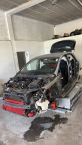 Voiture grise en cours de démontage sur un pont élévateur dans un garage. Moteur exposé, intercooler rouge visible, portes retirées. Flaque de liquide au sol. Panneau "DAE".